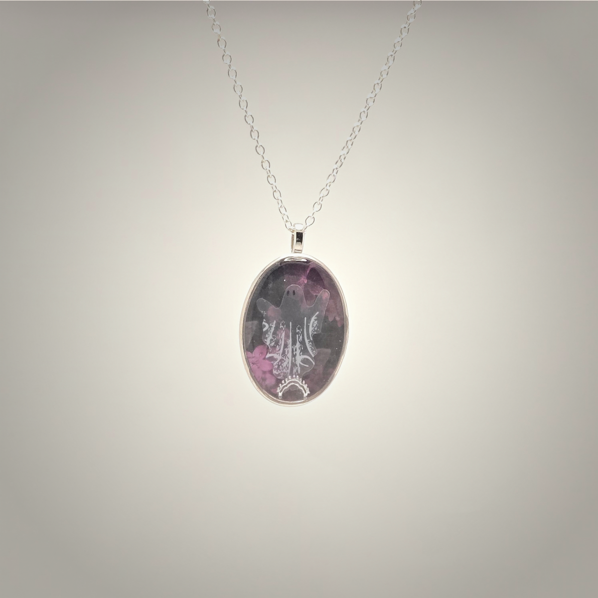 An oval pendant necklace featuring a purple and pink floral design with a silver-toned filigree pattern, suspended on a delicate silver chain against a neutral gray gradient background.