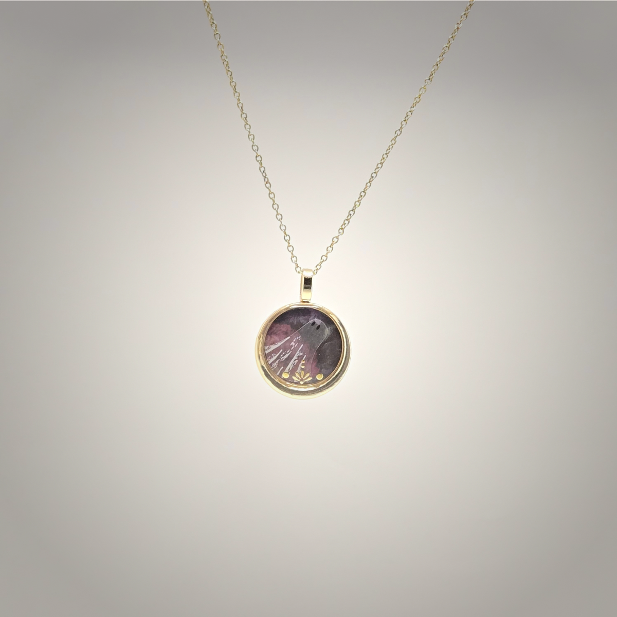 A gold chain necklace with a round pendant featuring a purple and pink feather design with tiny gold glitter flecks, displayed against a neutral gray gradient background.