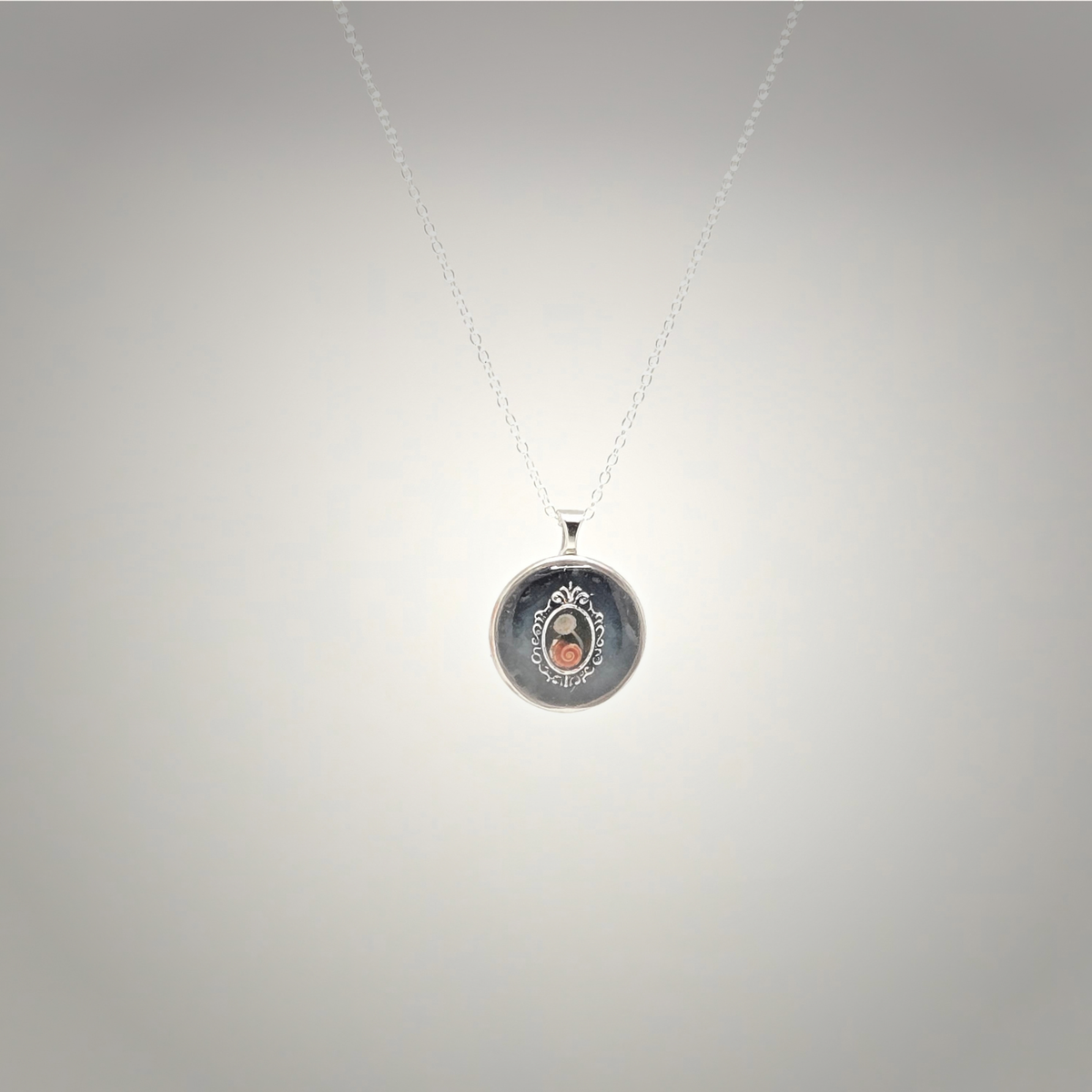 A silver pendant necklace featuring a circular medallion with a dark background, silver border detailing, and a small central rose gold oval accent, suspended from a delicate silver chain against a light gray gradient background.