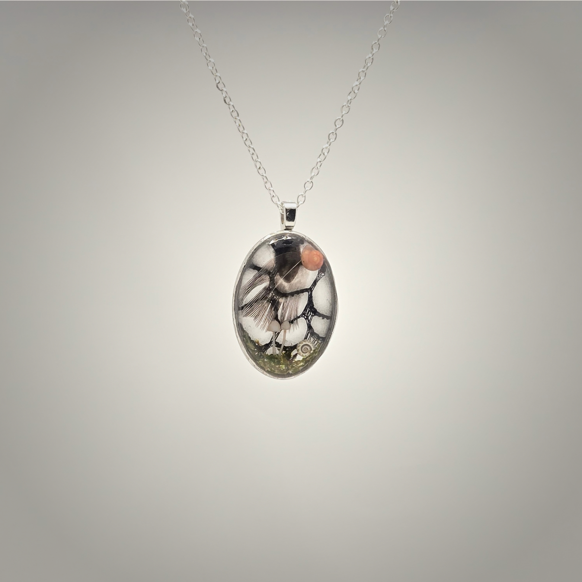 An oval pendant necklace featuring a detailed illustration of a bird in black, white, and brown tones with a small orange accent, suspended on a delicate silver chain against a neutral gray gradient background.
