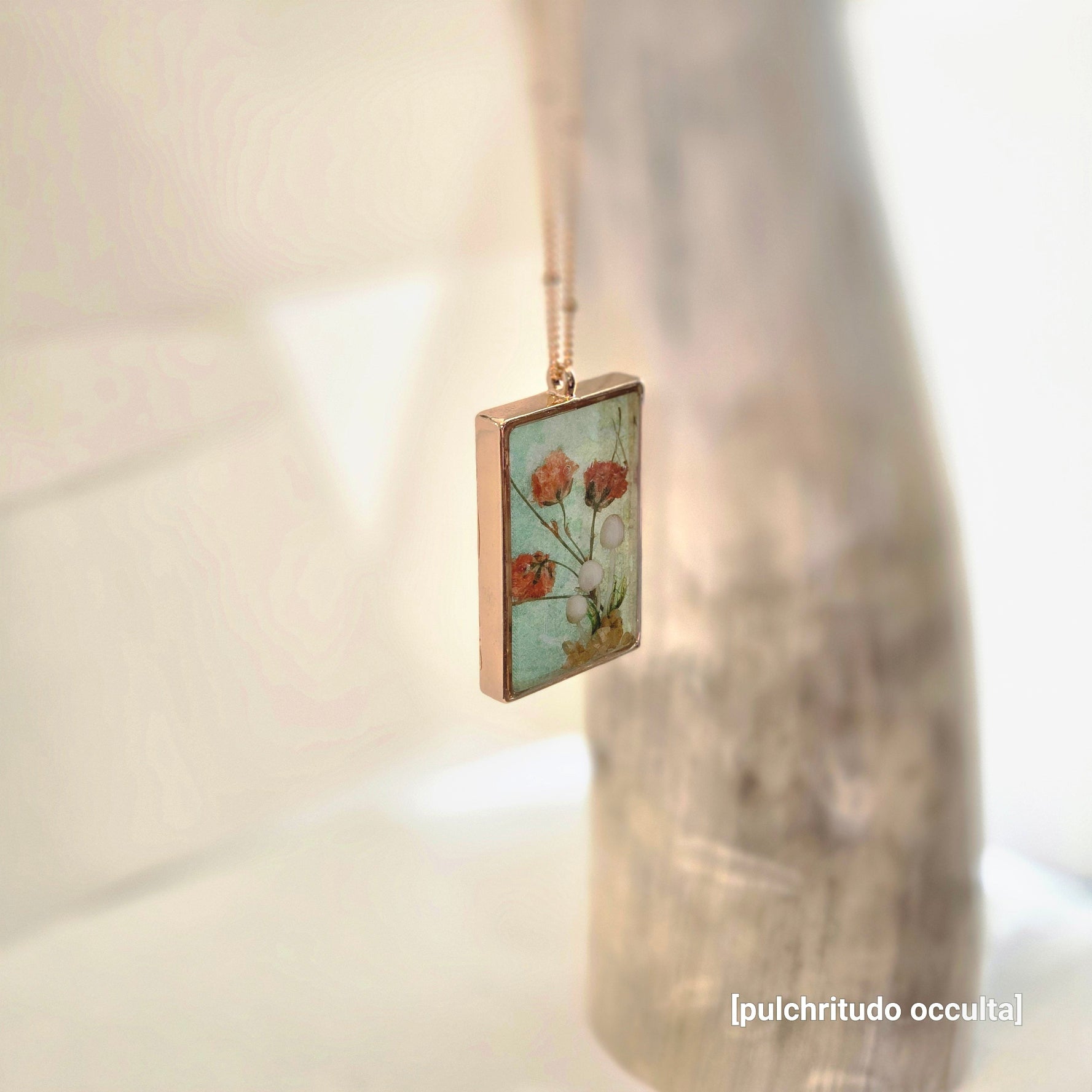 Square pendant with floral design on a chain against a blurred background