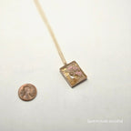 Decorative pick with floral design on a white background, next to a penny for scale.