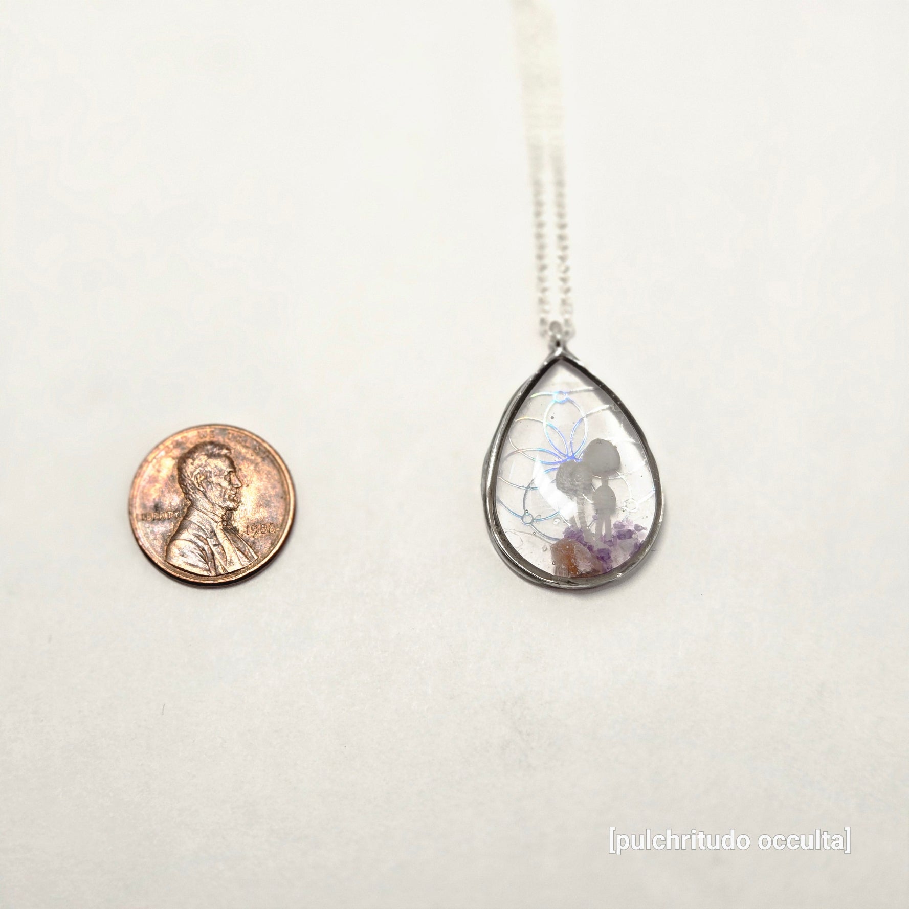 Teardrop-shaped pendant with a coin underneath on a white background