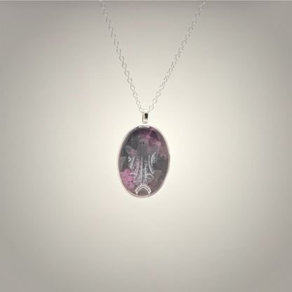 An oval pendant necklace featuring a purple and pink floral design with a silver-toned filigree pattern, suspended on a delicate silver chain against a neutral gray gradient background.