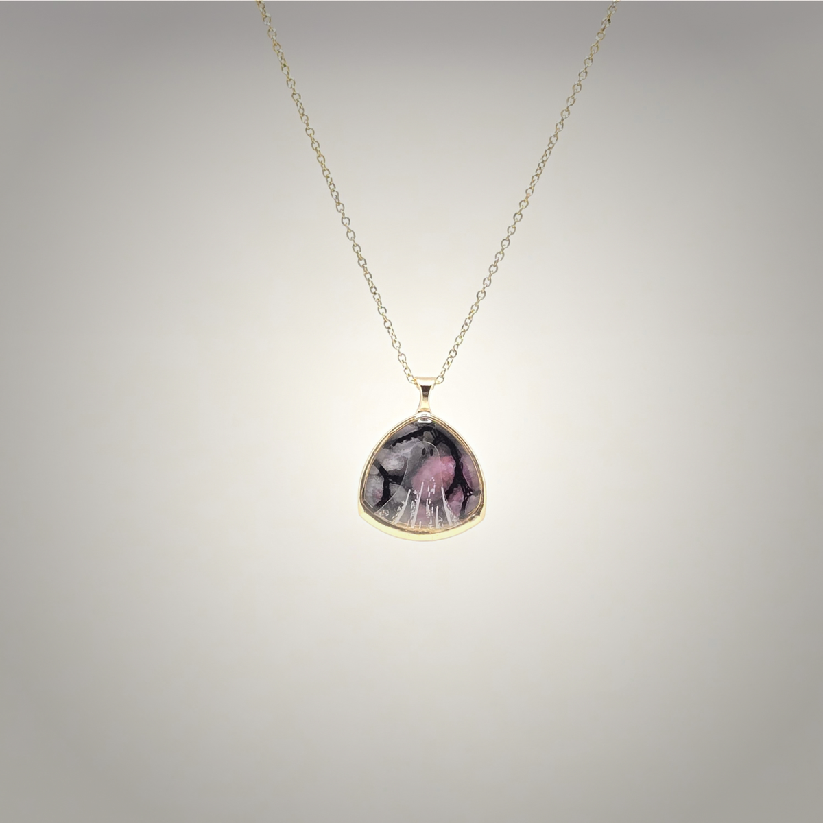 A gold chain necklace with a teardrop-shaped pendant featuring a pink and black marbled pattern, set in a gold bezel against a light gray gradient background.