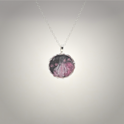 A silver chain necklace with a round, textured bezel pendant featuring a ghostly figure in a white robe surrounded by pink flowers on a dark background, shown against a neutral gray gradient background.