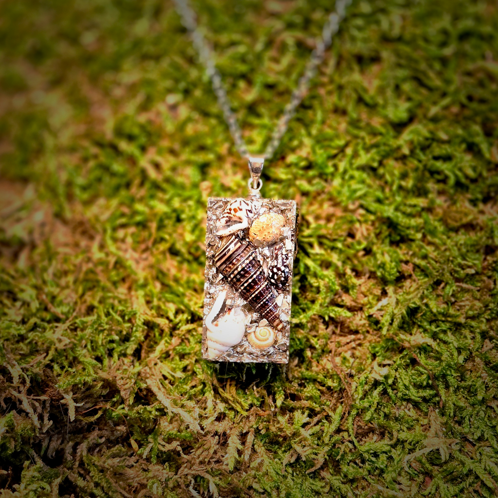A rectangular pendant necklace featuring various seashells and small natural inclusions set in a clear resin, hanging from a silver-toned chain on a bed of green moss.