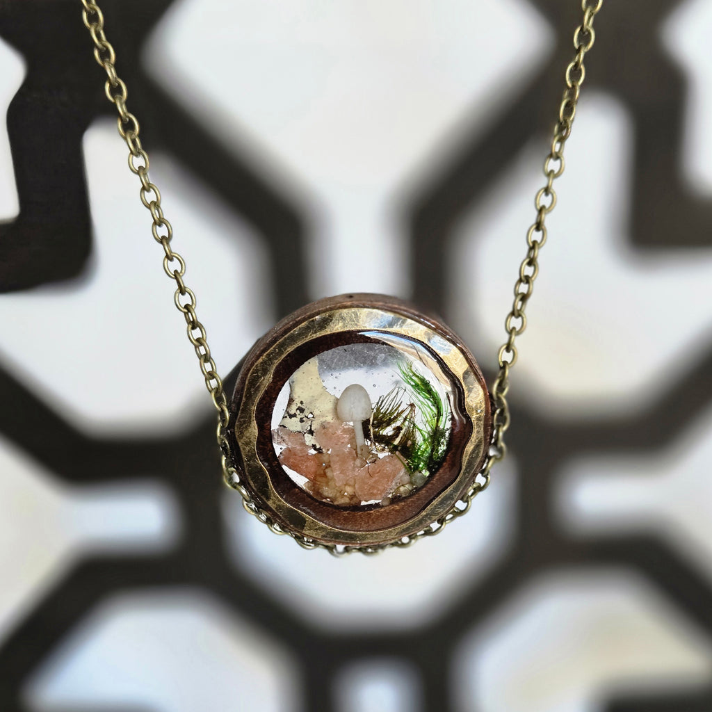 A round pendant necklace with a glass dome containing a tiny white mushroom, green moss, and small pink crystals, creating a miniature forest scene, hanging from a 24‑inch antique‑gold chain.