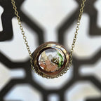 A round pendant necklace with a glass dome containing a tiny white mushroom, green moss, and small pink crystals, creating a miniature forest scene, hanging from a 24‑inch antique‑gold chain.