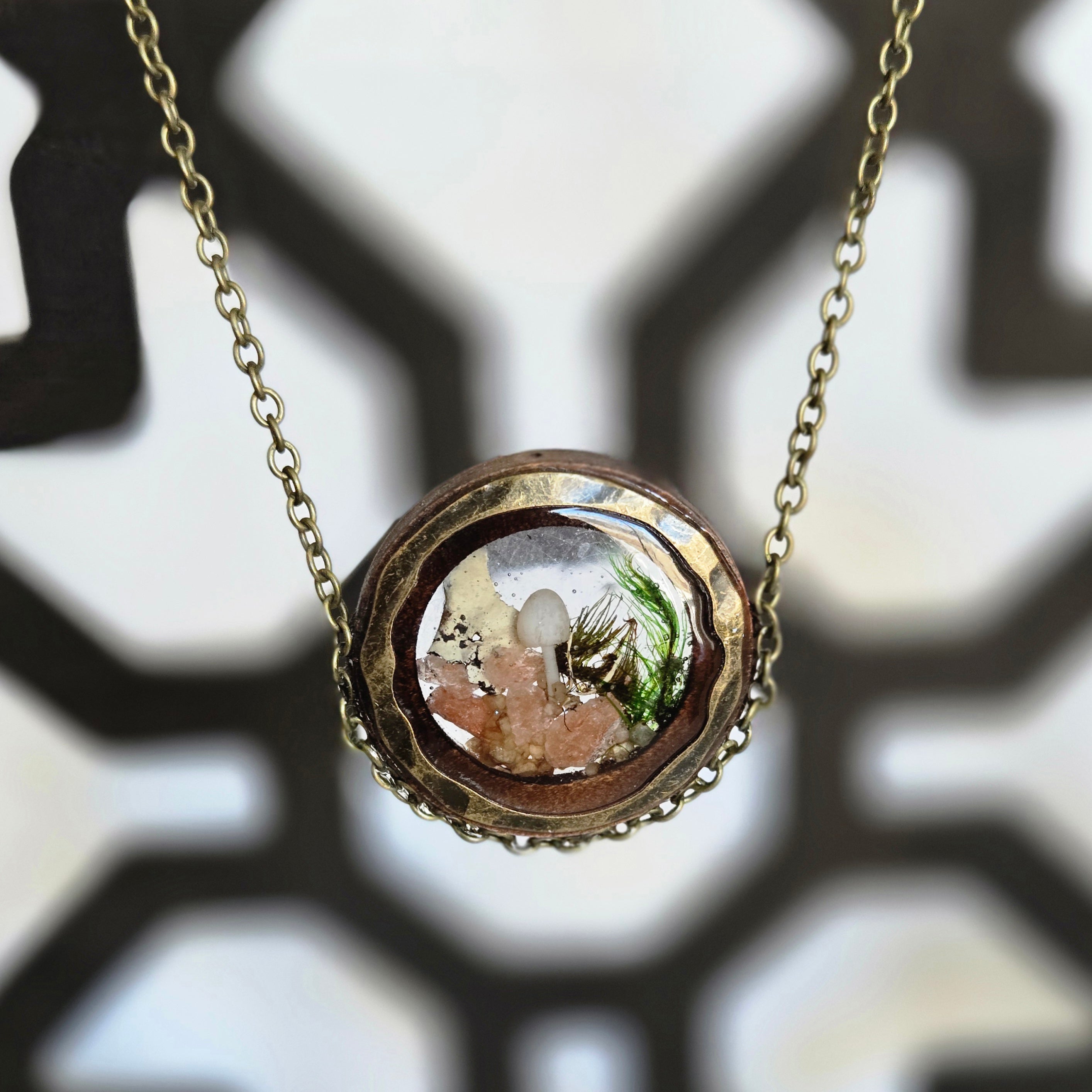 A round pendant necklace with a glass dome containing a tiny white mushroom, green moss, and small pink crystals, creating a miniature forest scene, hanging from a 24‑inch antique‑gold chain.