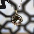 A round resin pendant necklace containing a tiny white mushroom, green moss, and small stones over a delicate lace‑leaf background, hanging from a 24‑inch antique‑gold chain.
