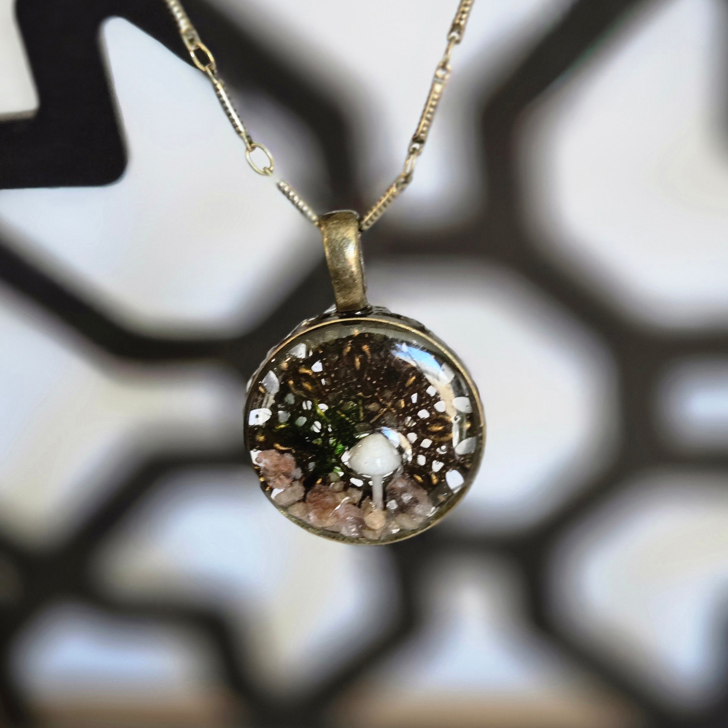 A round resin pendant necklace containing a tiny white mushroom, green moss, and small stones over a delicate lace‑leaf background, hanging from a 24‑inch antique‑gold chain.