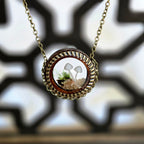 A round pendant necklace with a glass dome containing two tiny white mushrooms, green moss, and small beige stones, creating a miniature forest scene, hanging from a 22‑inch antique‑gold chain.