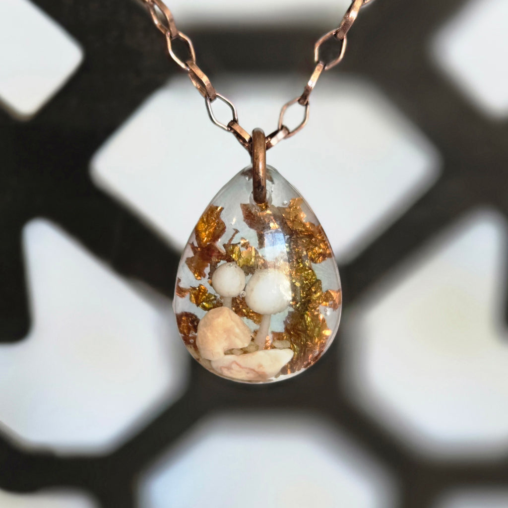 A teardrop resin pendant containing two small white mushrooms, beige botanicals, and warm copper flakes, hanging from an 18‑inch antique‑gold chain.