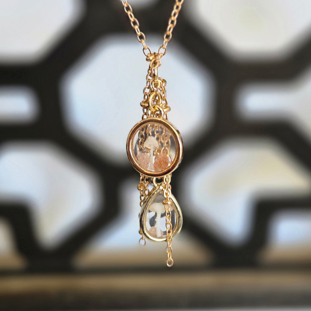 A gold‑tone necklace with two connected resin pendants—one round and one teardrop—each containing tiny mushrooms and botanical elements, hanging from a 20‑inch chain.