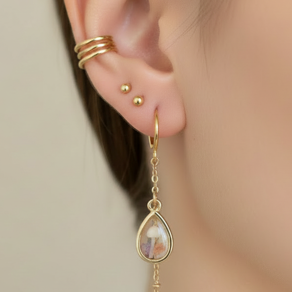 Gold earring with a teardrop-shaped pendant worn by a person against a neutral background