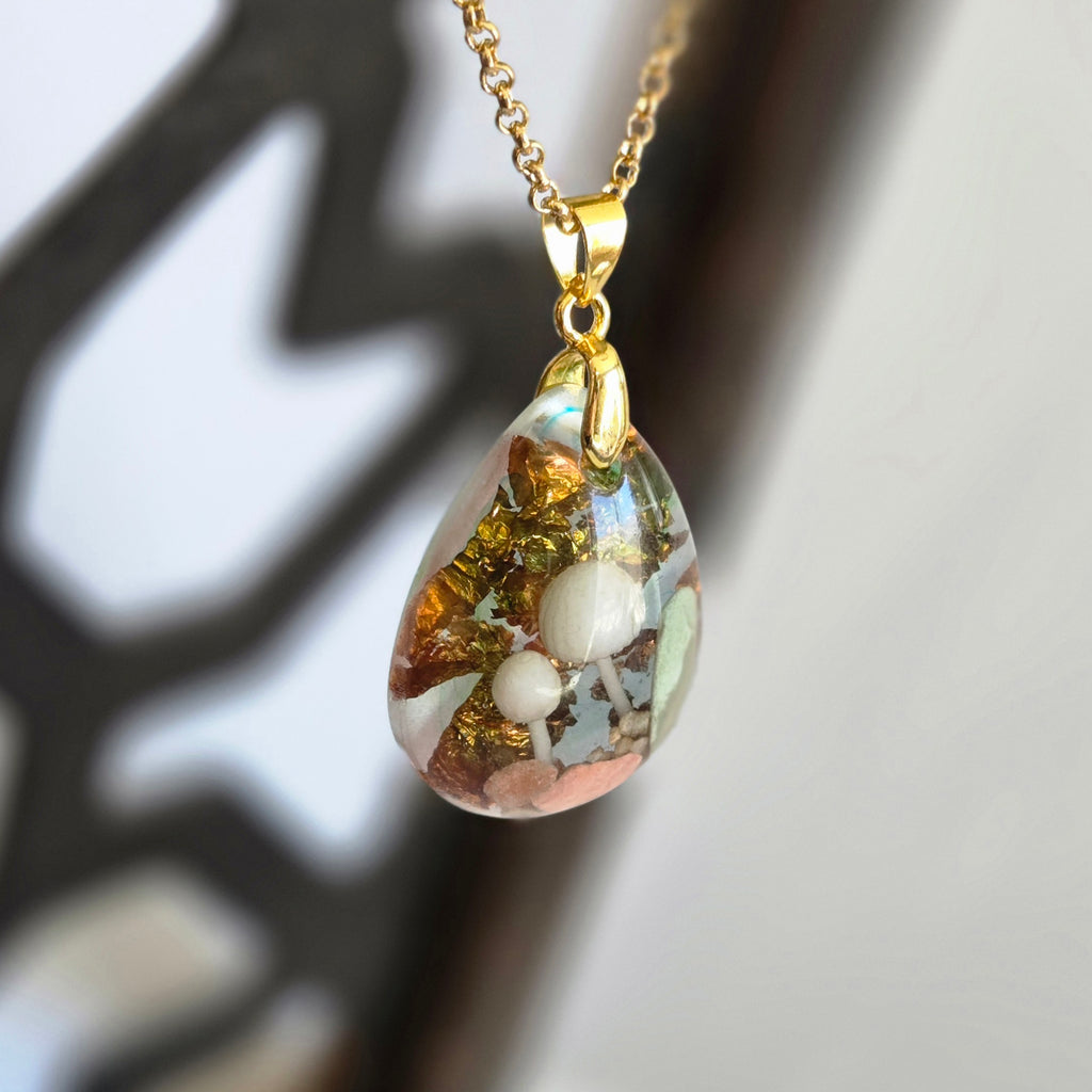 Teardrop-shaped pendant with embedded pearls on a gold chain against a blurred natural background