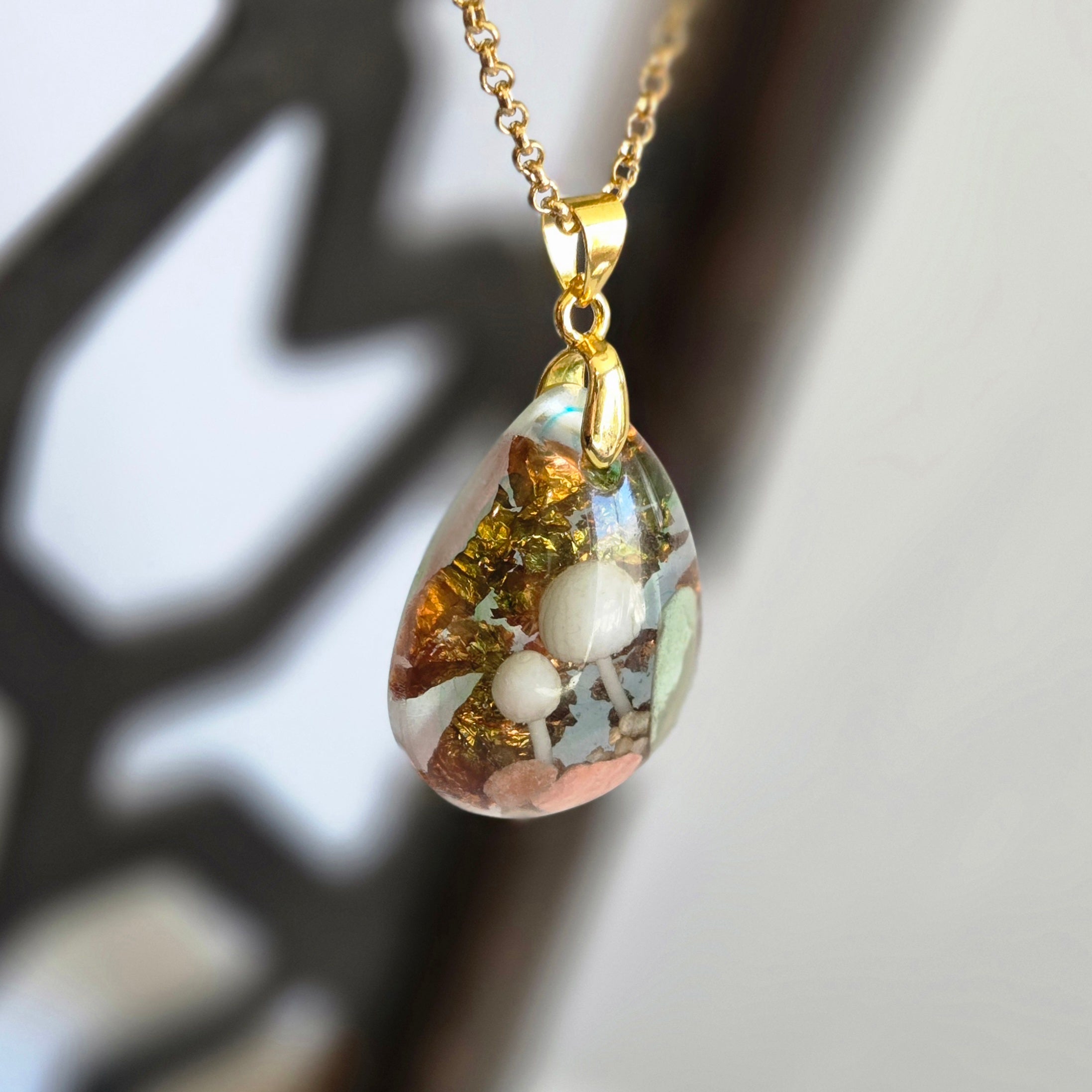 Teardrop-shaped pendant with embedded pearls on a gold chain against a blurred natural background