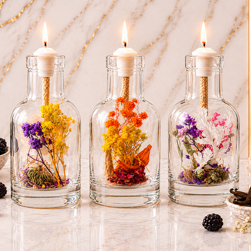 Three clear glass bottles with white pillar candles and dried flower arrangements, displayed on a marble surface with decorative blackberries and flowers against a marble wall.