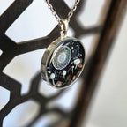 Necklace with a round pendant featuring a black background with white and gray elements, hanging against a blurred background.